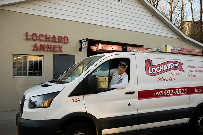 Lochard HVAC, Plumbing & Service branded vehicle in front of storefront with technician smiling, representing HVAC and plumbing maintenance in Sidney, OH including furnace tune-ups, AC maintenance, refrigerant checks, compressor diagnostics, plumbing inspections, leak detection, drain cleaning, water heater maintenance, and preventative home comfort system service.